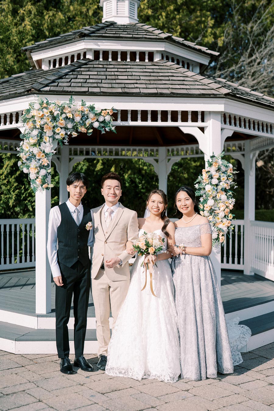 Wedding party posing in front of a flower-adorned gazebo. The bride in a lace gown holds a bouquet, standing beside the groom in a beige suit. Two bridesmaids join them, dressed in elegant dresses, all smiling in a serene garden setting.