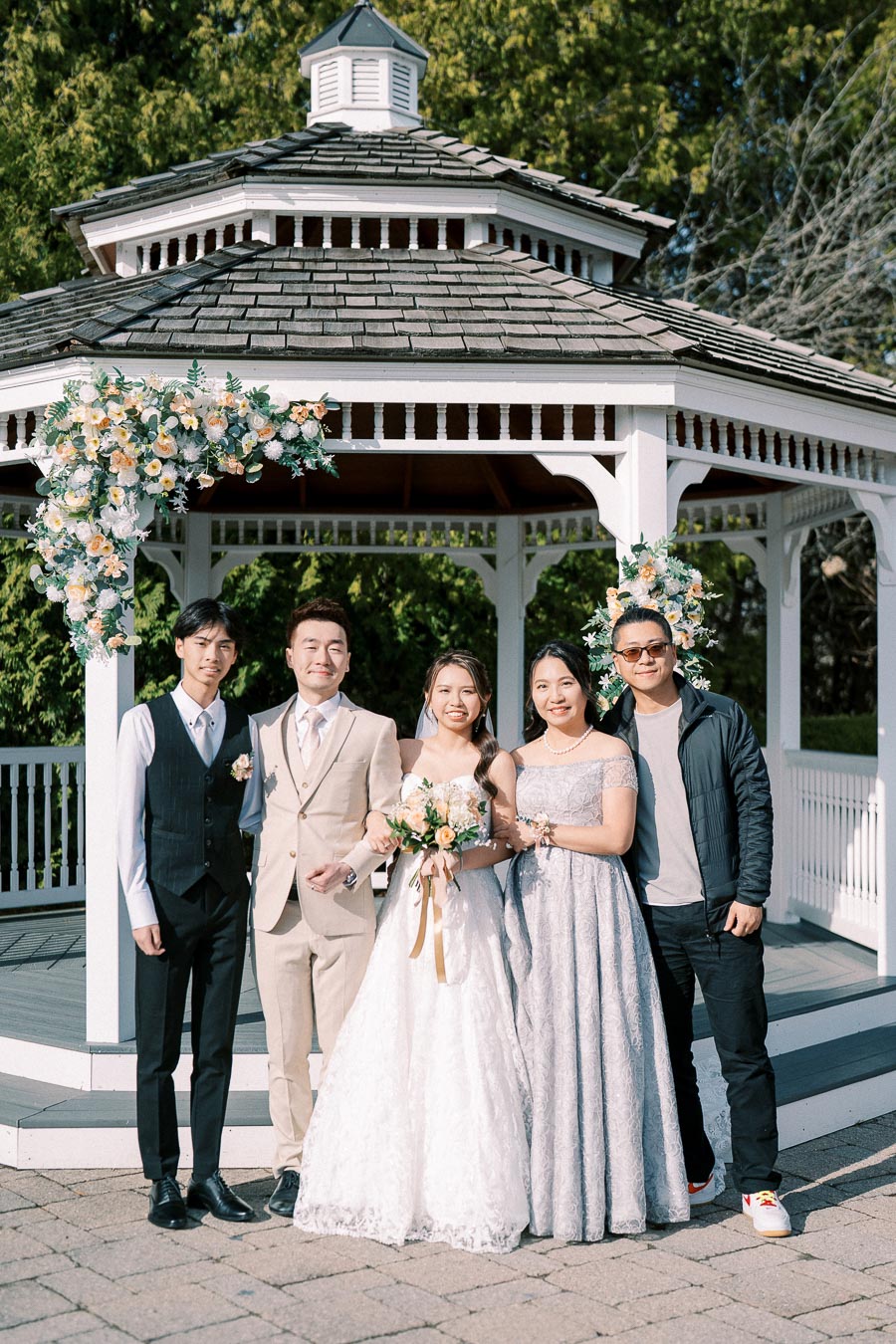 A group photo of five people standing together in front of a white gazebo decorated with flowers, including the bride in a white lace gown and groom in a beige suit, during an outdoor wedding ceremony.