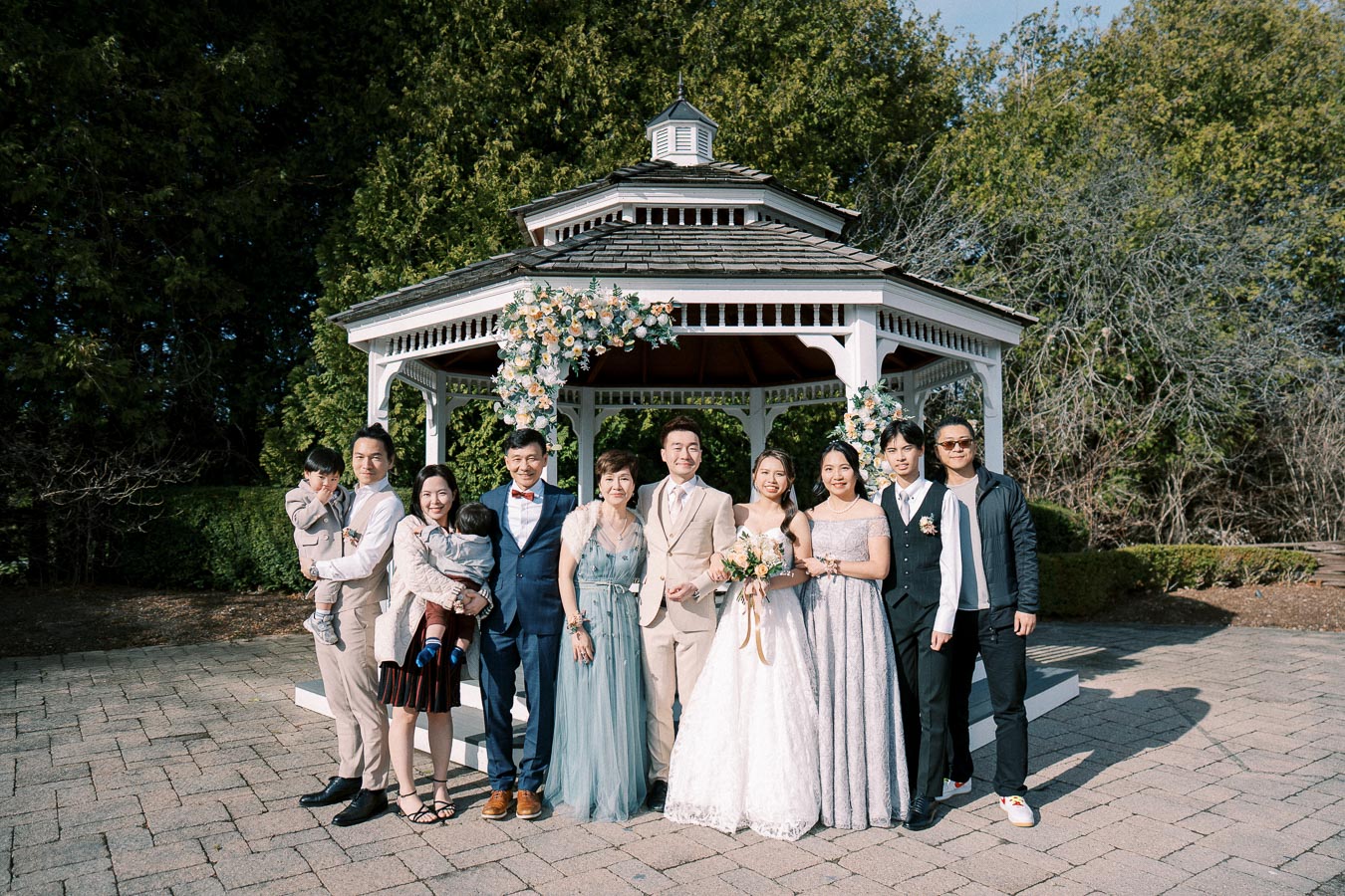 A family poses together in front of a floral-decorated white gazebo on a sunny day, showcasing a wedding celebration.