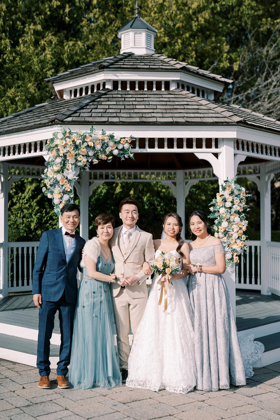 A happy couple poses with family members in elegant attire in front of a beautifully decorated gazebo, surrounded by lush greenery and floral arrangements, during a sunny outdoor wedding ceremony.