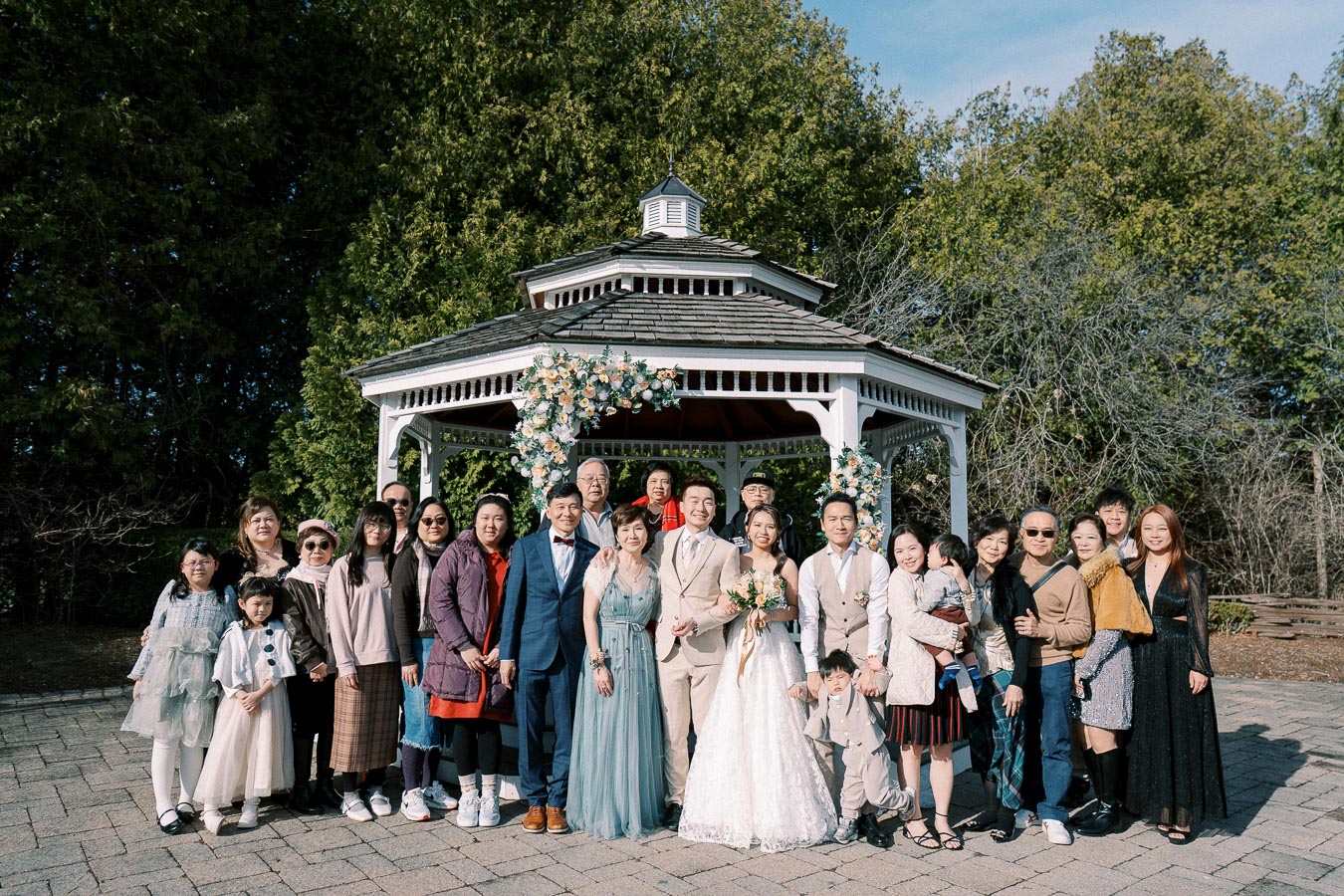 A large group of people smiling in front of a decorated gazebo at an outdoor wedding, surrounded by greenery and a clear sky.