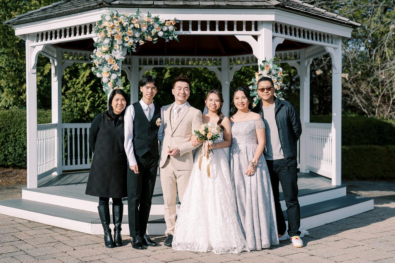 A group of six people pose in front of a white gazebo decorated with flowers. The bride and groom stand in the center, surrounded by family and friends, all dressed in formal attire. The scene is set outdoors on a sunny day, capturing a joyful wedding celebration.