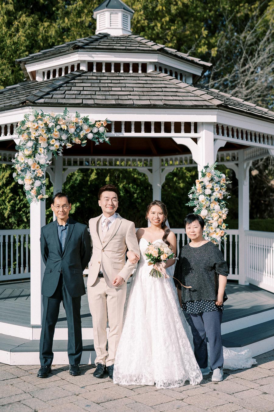 Wedding couple poses with family in front of a floral-decorated gazebo, outdoor ceremony setting.