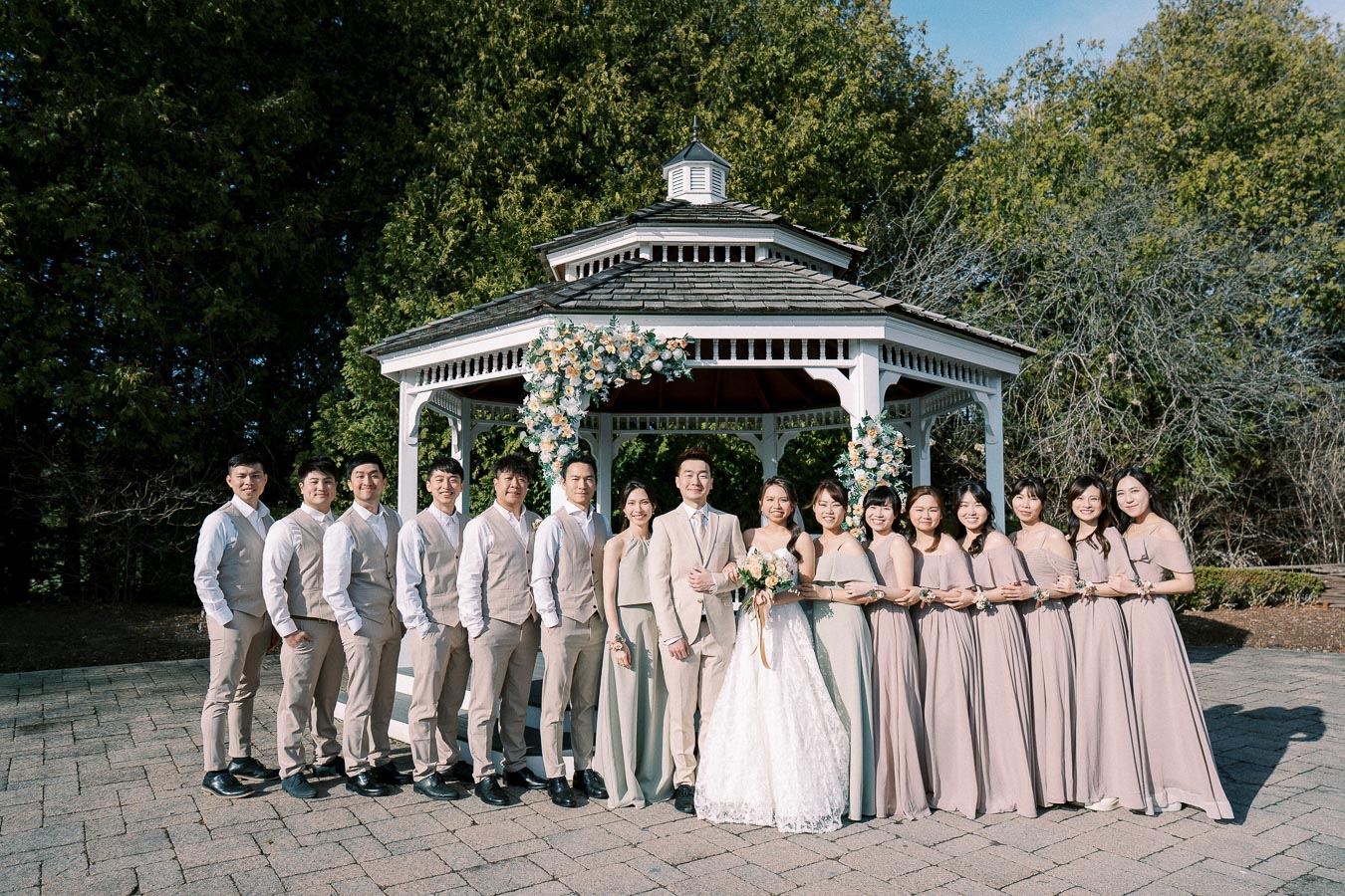 Wedding party posing in front of a decorated gazebo, featuring the bride and groom surrounded by bridesmaids in matching dresses and groomsmen in coordinated suits, set in a scenic outdoor venue.