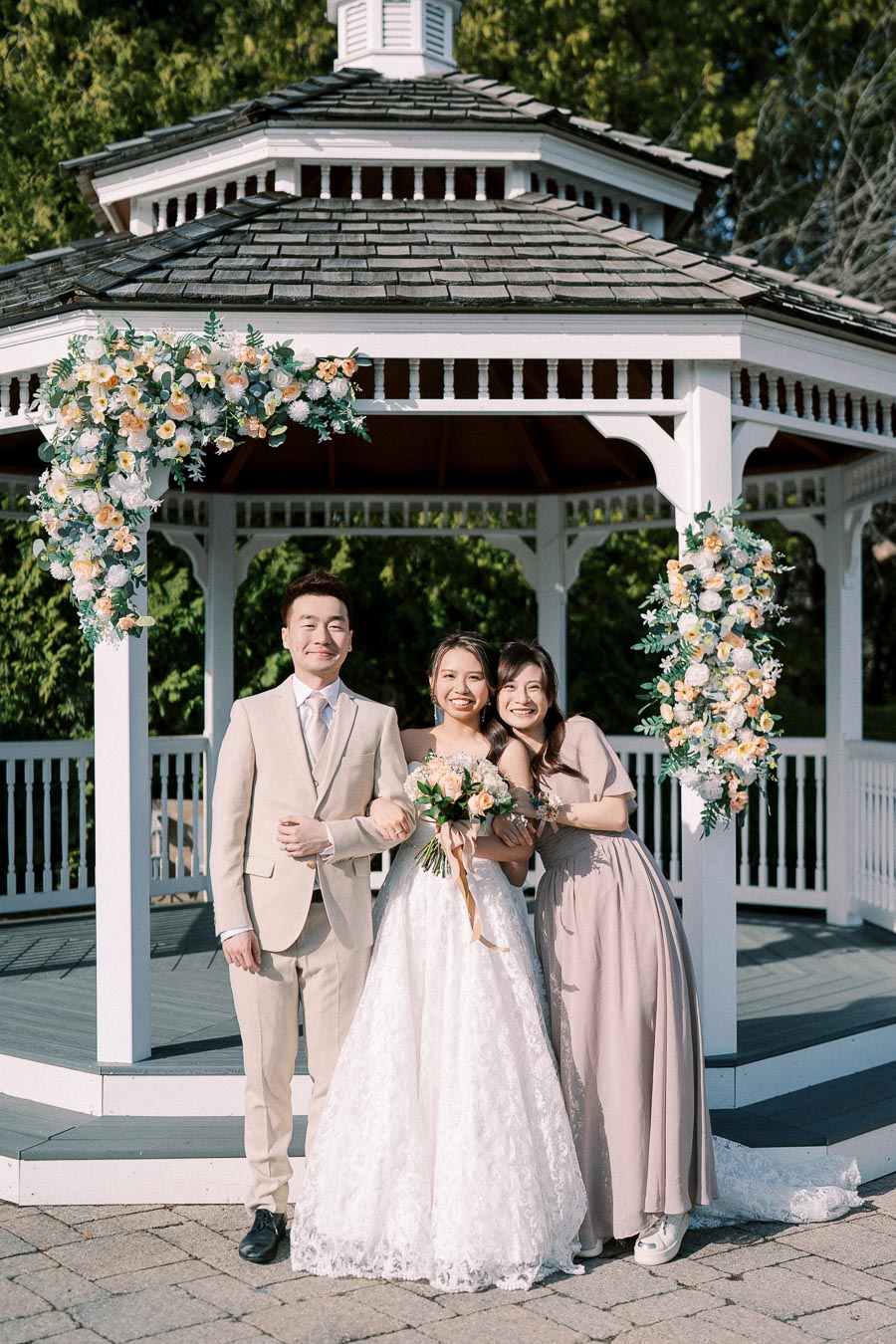 Elegant outdoor wedding ceremony with a smiling bride in a lace gown, standing between her groom in a beige suit and a bridesmaid in a taupe dress, in front of a beautifully decorated white gazebo adorned with fresh flowers.