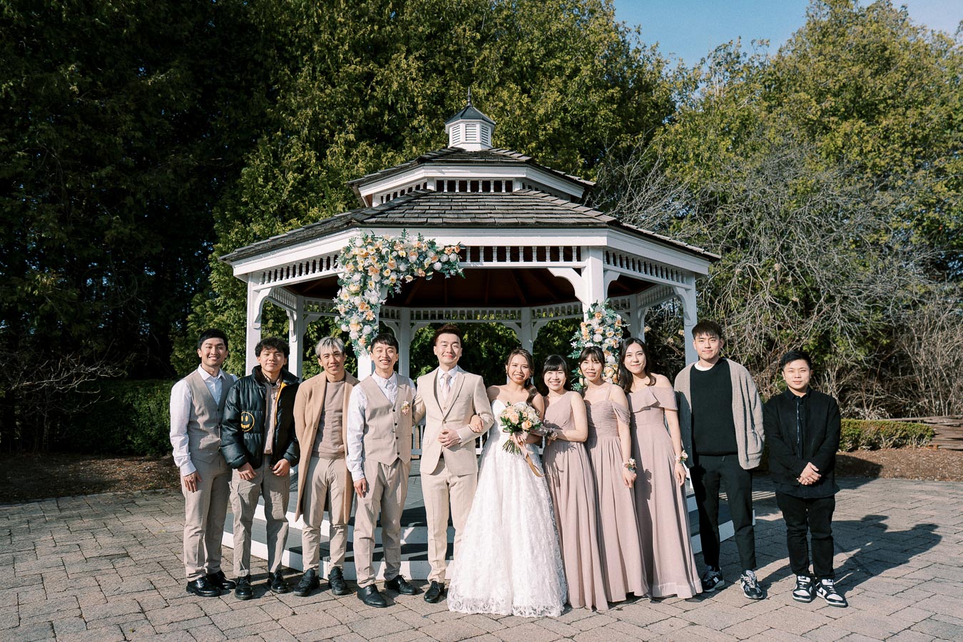 Wedding party posing in front of a decorated gazebo with lush greenery in the background. The bride is in a white gown holding a bouquet, surrounded by the groom and attendants in coordinated attire under a clear blue sky.