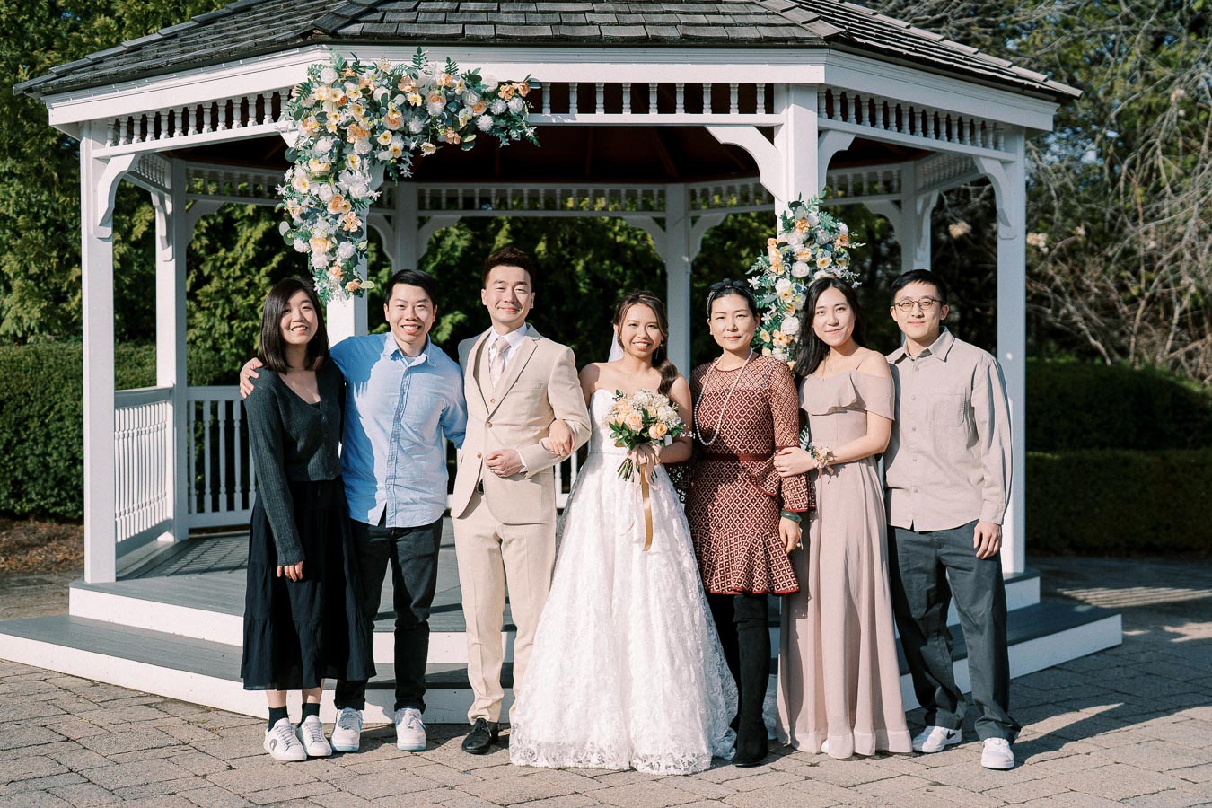 A group of people standing together posing for a wedding photo in front of a decorated gazebo. A bride in a white dress holds a bouquet, accompanied by others dressed in formal attire, against a backdrop of greenery.