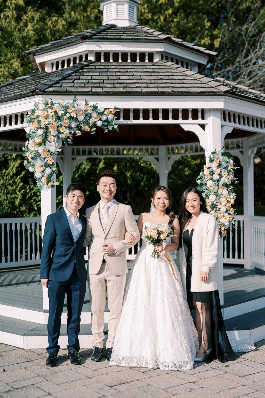 A bride and groom standing with two guests in formal attire in front of a decorated gazebo with flowers on a sunny day.