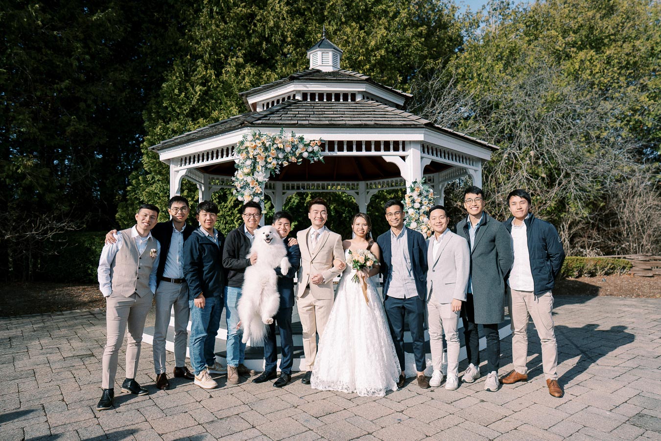 A wedding party poses in front of a white gazebo adorned with flowers, featuring the bride in a white gown, the groom in a beige suit, several groomsmen, and a fluffy white dog.