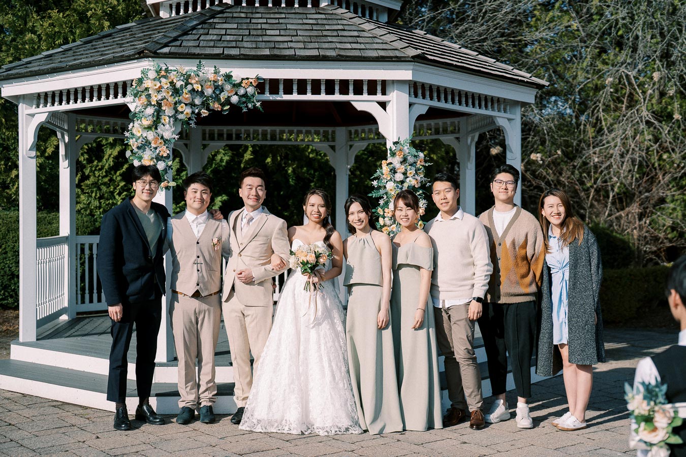 Wedding party posing in front of a decorated gazebo, featuring a bride in a white gown and groom in a beige suit, surrounded by bridesmaids and groomsmen under a clear blue sky.