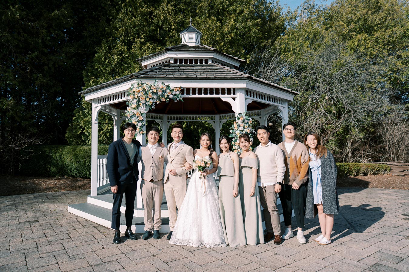 Wedding party standing in front of a decorated gazebo in a garden, featuring the bride in a white gown, the groom in a beige suit, and bridesmaids in matching dresses, with groomsmen and guests smiling in casual attire.