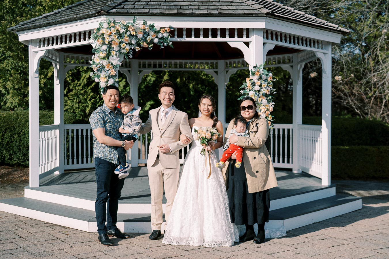 A happy wedding couple poses with family members in front of a beautifully decorated gazebo on a sunny day. The bride, dressed in an elegant white gown, holds a bouquet, while the groom stands beside her in a light beige suit.