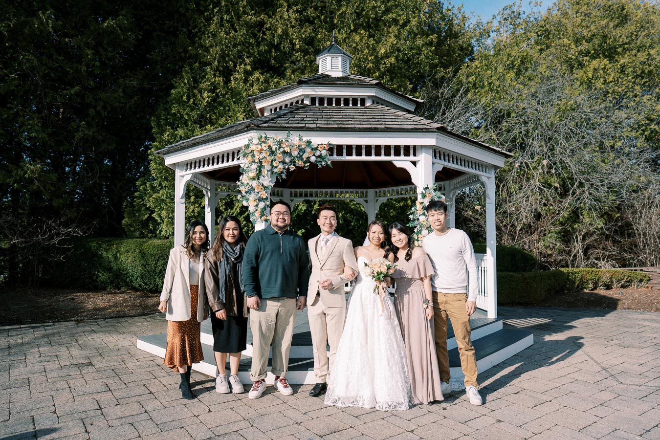 A joyful wedding group photo featuring a smiling bridal couple and guests standing in front of a white gazebo decorated with flowers, set in a lush garden backdrop.