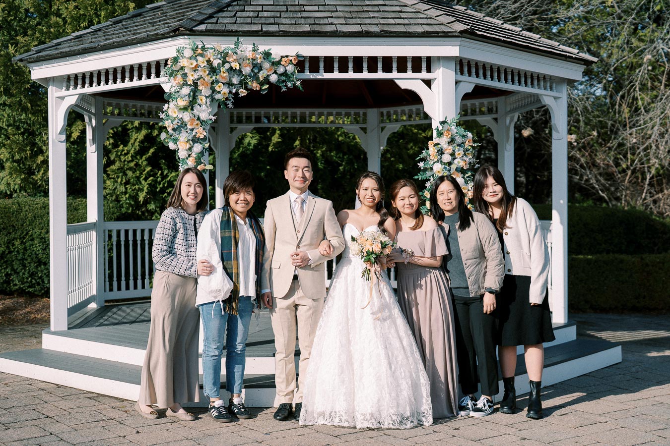 Wedding party posing in front of a decorated gazebo, featuring a bride in a white gown holding a bouquet, standing beside the groom in a beige suit, surrounded by smiling family members and friends on a sunny day.