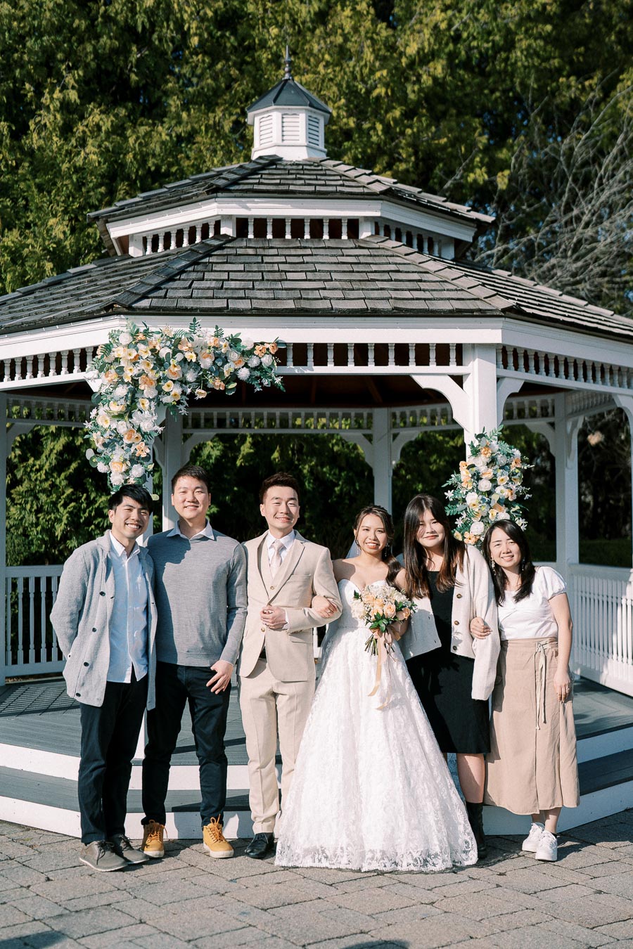 A group poses in front of a decorated gazebo at an outdoor wedding. The bride in a white gown and the groom in a beige suit stand in the center, surrounded by smiling friends. Lush greenery provides a scenic backdrop.