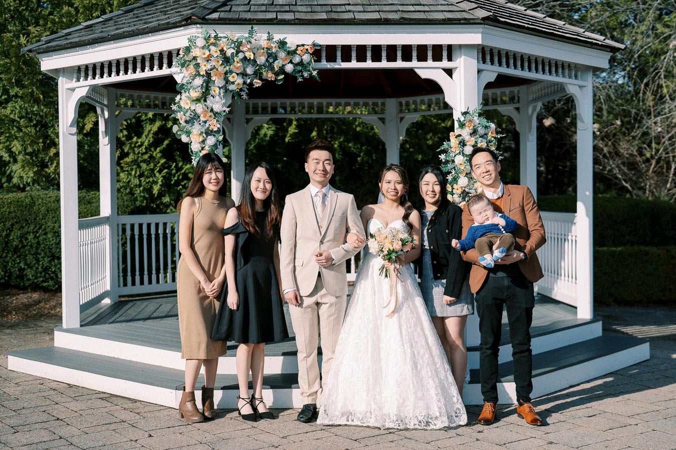 A wedding group poses in front of a decorated white gazebo, featuring the bride in a lace gown, the groom in a light suit, and four guests smiling, with one holding a baby. The gazebo is adorned with floral arrangements, adding to the picturesque outdoor setting.