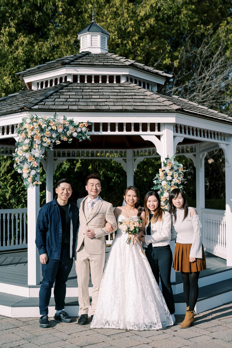 A joyful wedding group poses in front of a beautifully decorated gazebo. The bride, wearing a lace wedding gown, holds a bouquet of flowers, while the groom stands beside her in a beige suit. They are surrounded by three friends under a clear blue sky, with lush greenery in the background.