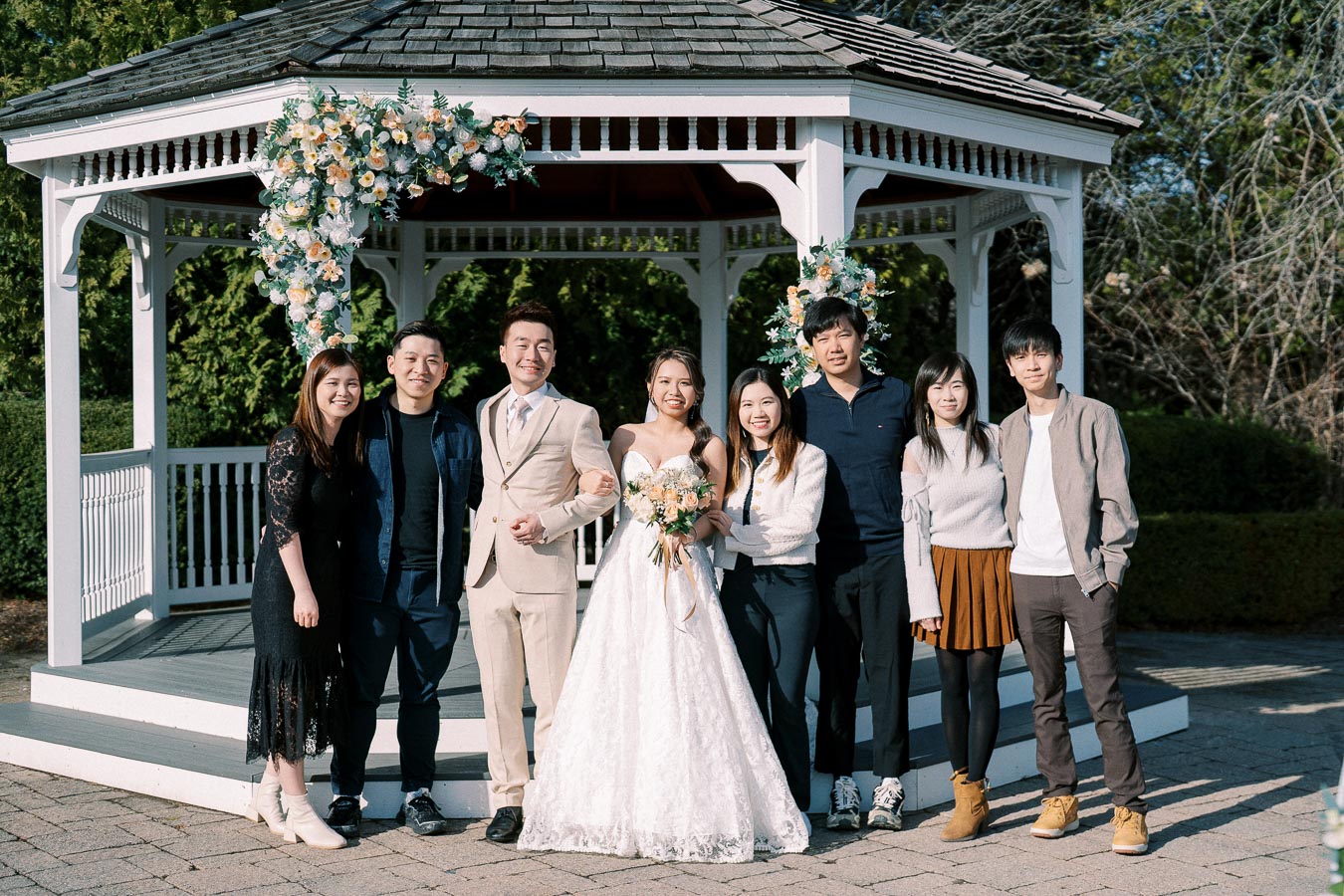 A group photo taken in front of a white gazebo decorated with flower arrangements, featuring a bride in a white gown holding a bouquet, a groom in a beige suit, and family or friends casually dressed, all smiling and standing on a paved path in a lush garden setting.