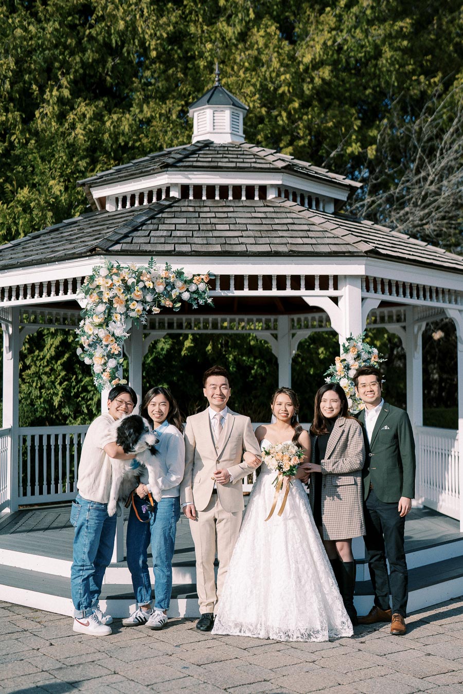 A group of people posing in front of a decorated gazebo during a wedding. The bride and groom stand in the center, smiling happily. The bride is in a white dress holding a bouquet, while the groom is in a beige suit. Friends and family surround them, including someone holding a black and white dog. The gazebo is adorned with flowers, and