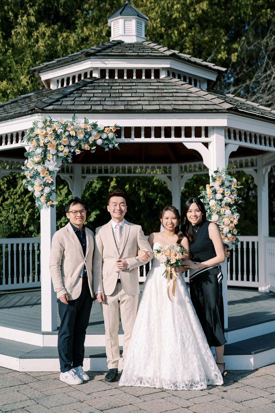 A bride and groom in wedding attire smiling with two guests posing in front of a decorated white gazebo on a sunny day.