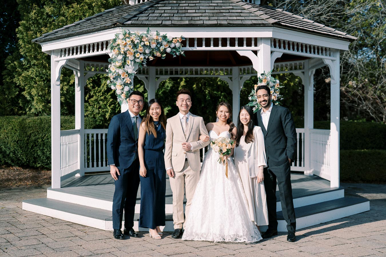 Group posing at an outdoor wedding by a decorated gazebo with floral arrangements, bride in white gown holding bouquet, groom in beige suit, surrounded by friends in formal attire.