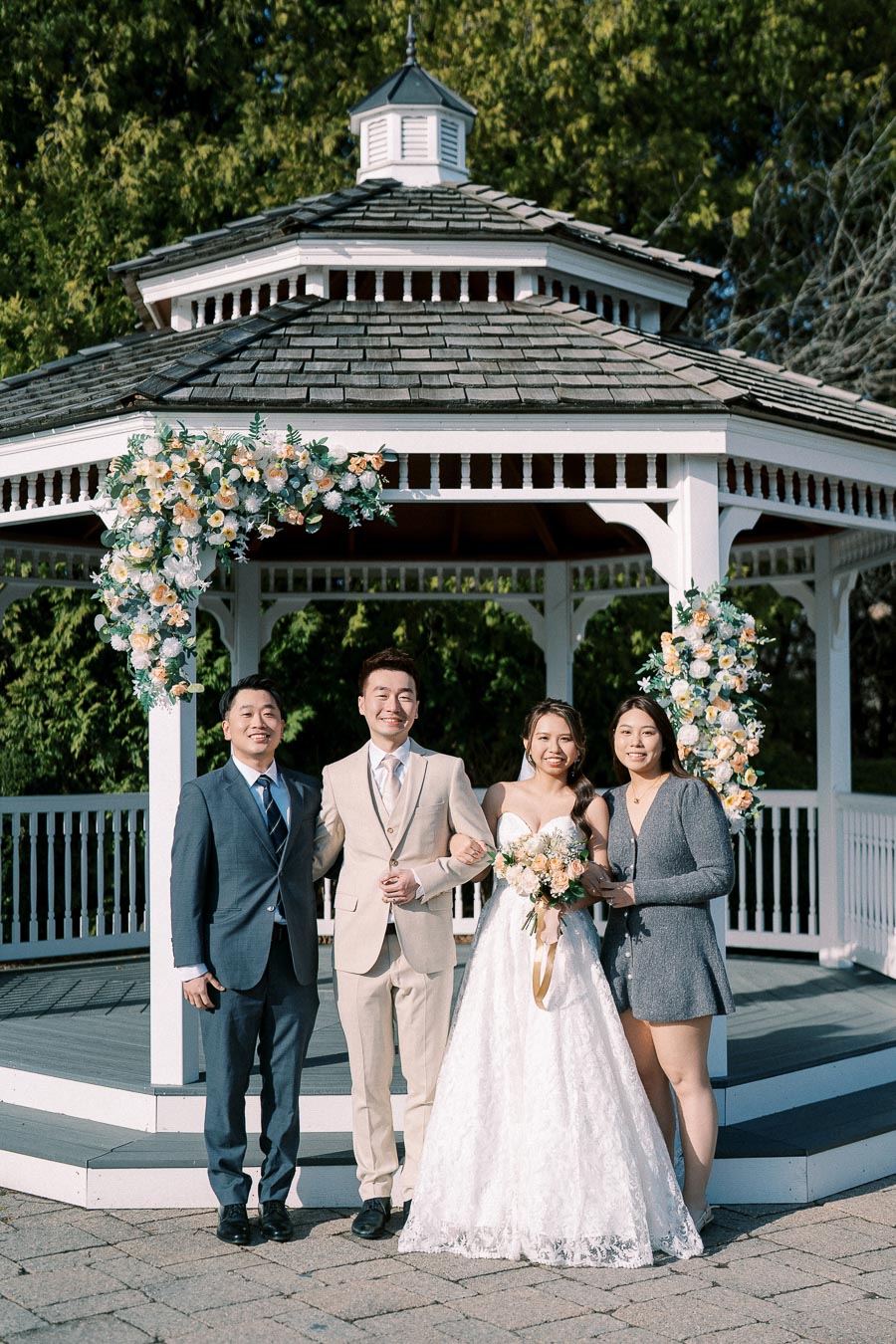 A bride and groom posing with two guests in formal attire in front of a floral-decorated gazebo, outdoors on a sunny day.