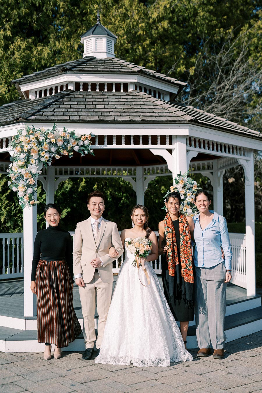 A happy wedding group poses in front of a floral-decorated white gazebo. The bride holds a bouquet, wearing a lace gown, while standing next to the groom in a light beige suit. Two women and another person stand beside them, completing the joyful outdoor celebration.
