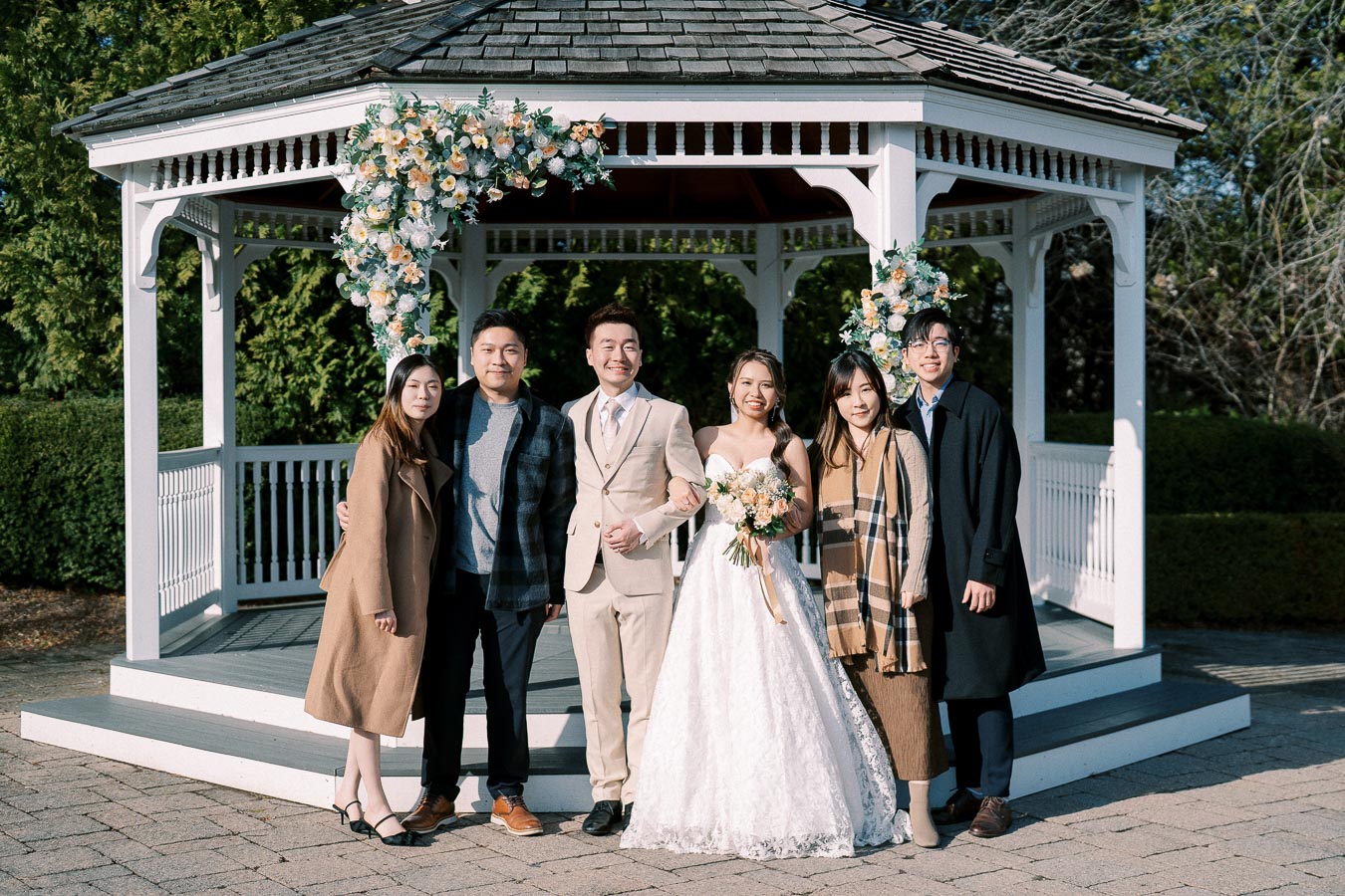A wedding couple stands with friends in front of a decorative gazebo adorned with floral arrangements, surrounded by greenery on a sunny day.