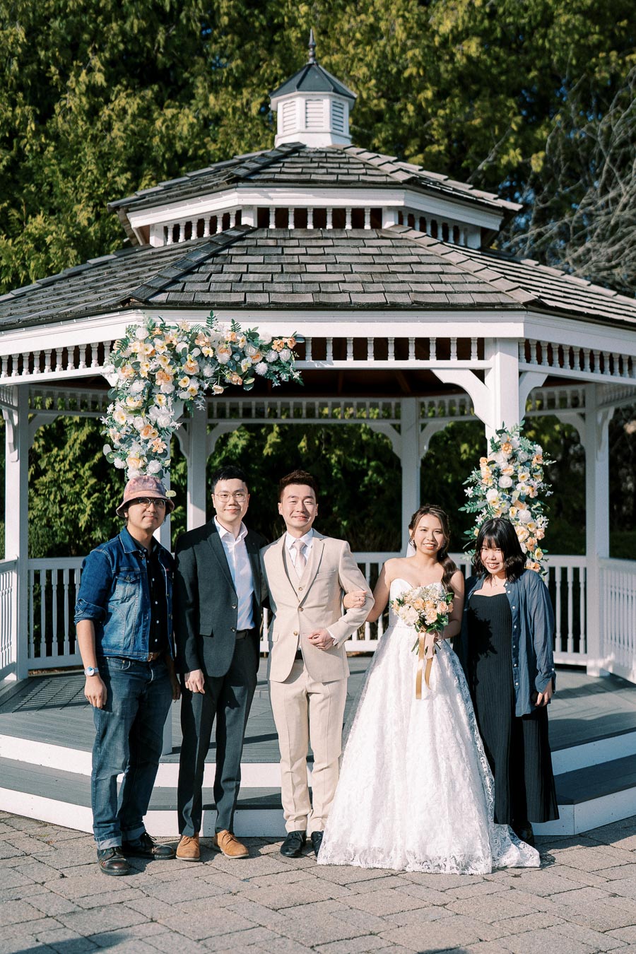 Wedding celebration at a floral-decorated gazebo with a bride in a white dress holding a bouquet, groom in a beige suit, and three guests posing together on a sunny day.