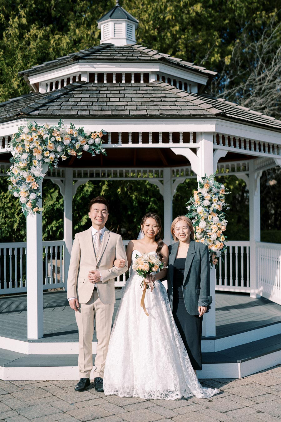 A bride in a white lace dress and groom in a beige suit standing with a woman in a dark outfit, smiling in front of a flower-adorned gazebo.