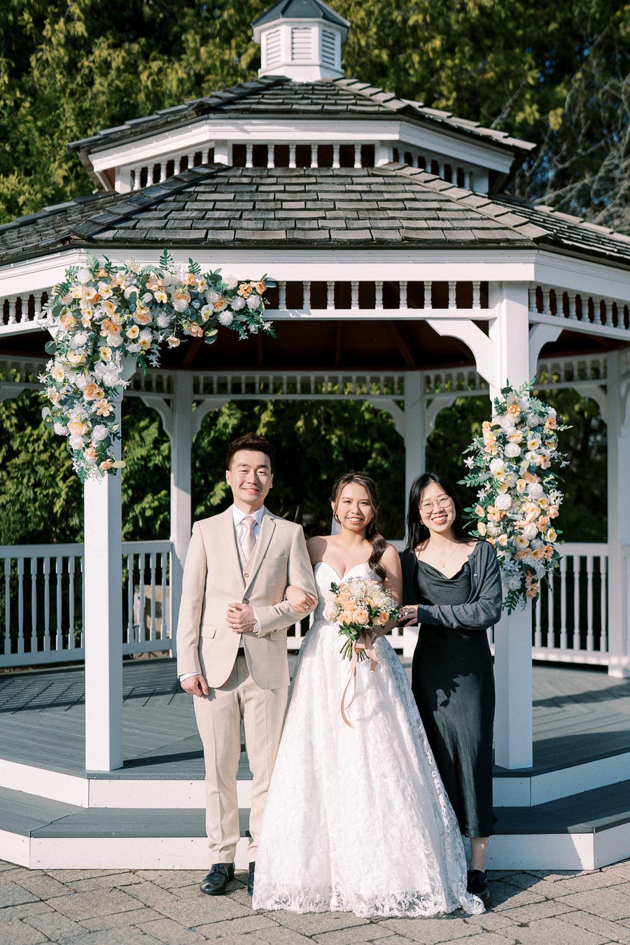 A bride and groom posing with a guest in front of a floral-decorated wooden gazebo, surrounded by lush greenery, on a sunny wedding day.