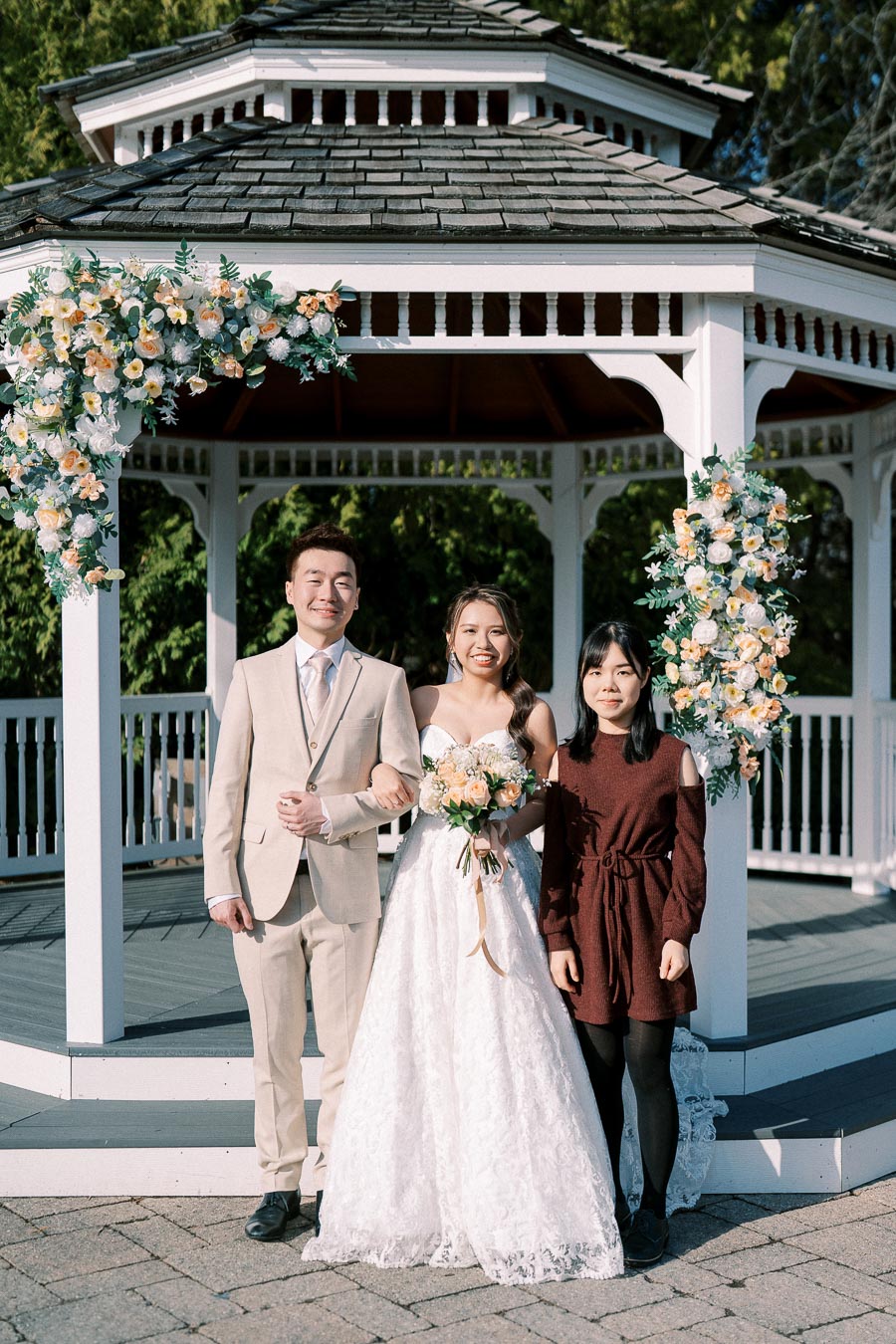 Wedding couple posing with a guest in front of a floral-decorated gazebo on a sunny day.