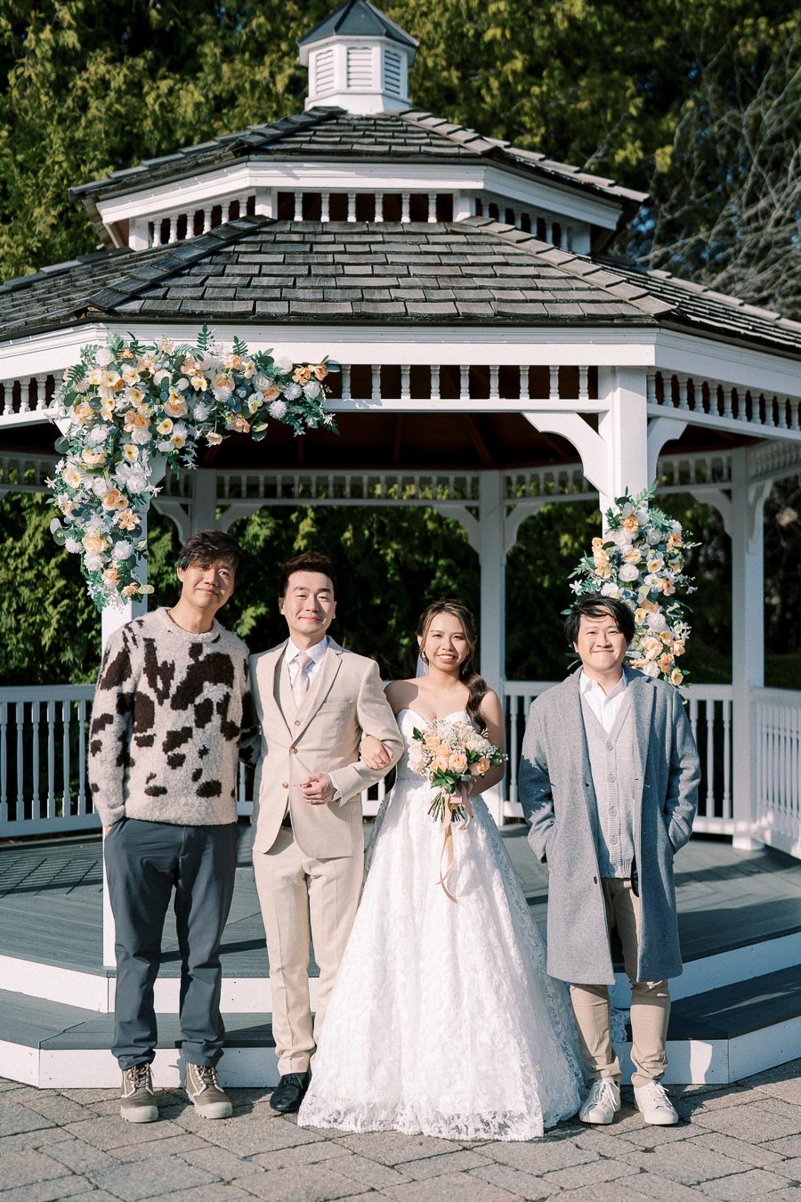 A happy couple in wedding attire poses with two guests in front of a floral-decorated gazebo on a sunny day.