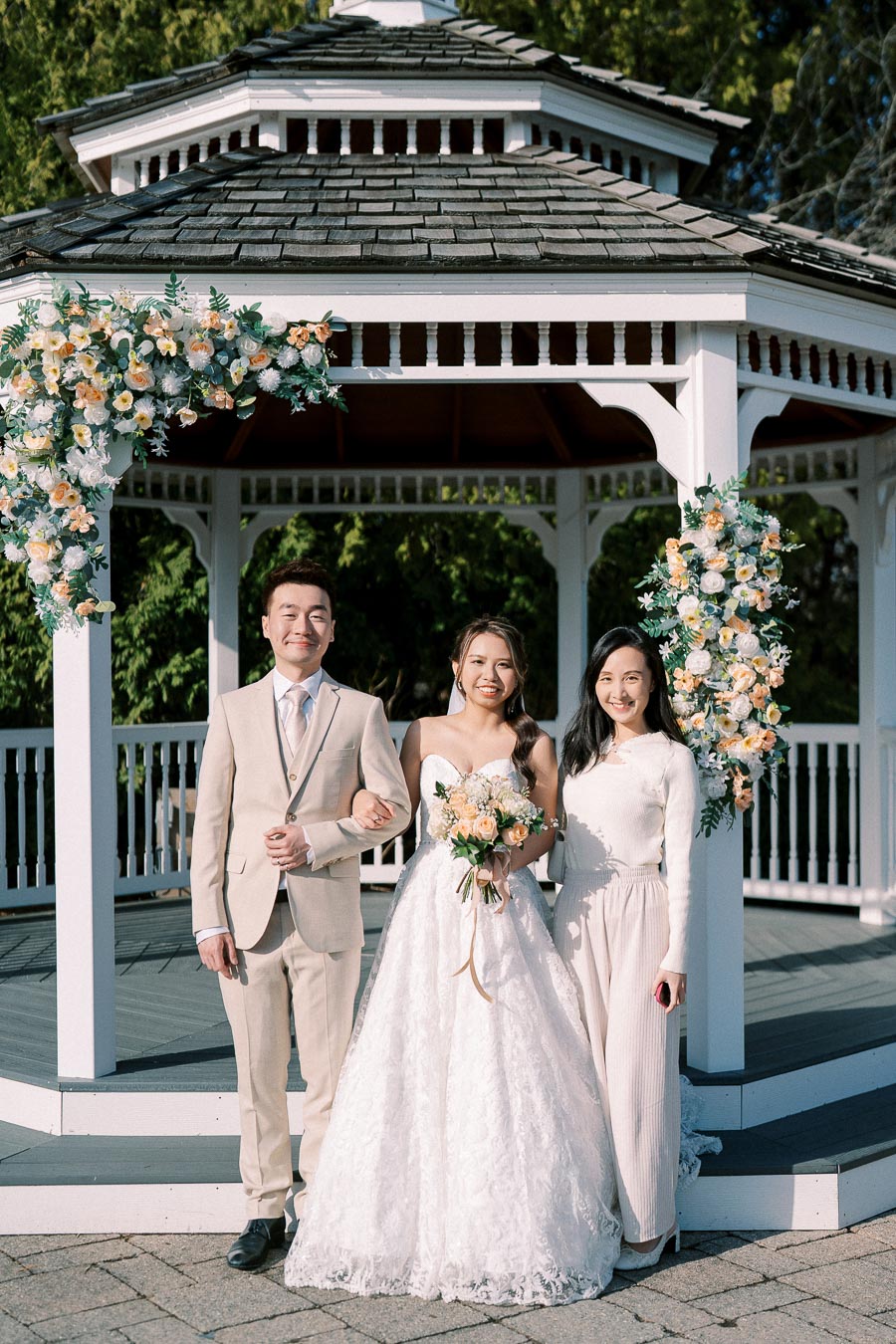 A bride in a white lace gown holding a bouquet, standing with two people in formal attire in front of a flower-adorned gazebo. Bright and sunny outdoor wedding scene with greenery in the background.