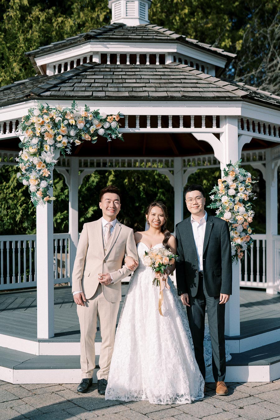 Bride and groom standing with a friend in front of a beautifully decorated gazebo adorned with floral arrangements, set against a lush green backdrop at an outdoor wedding ceremony.
