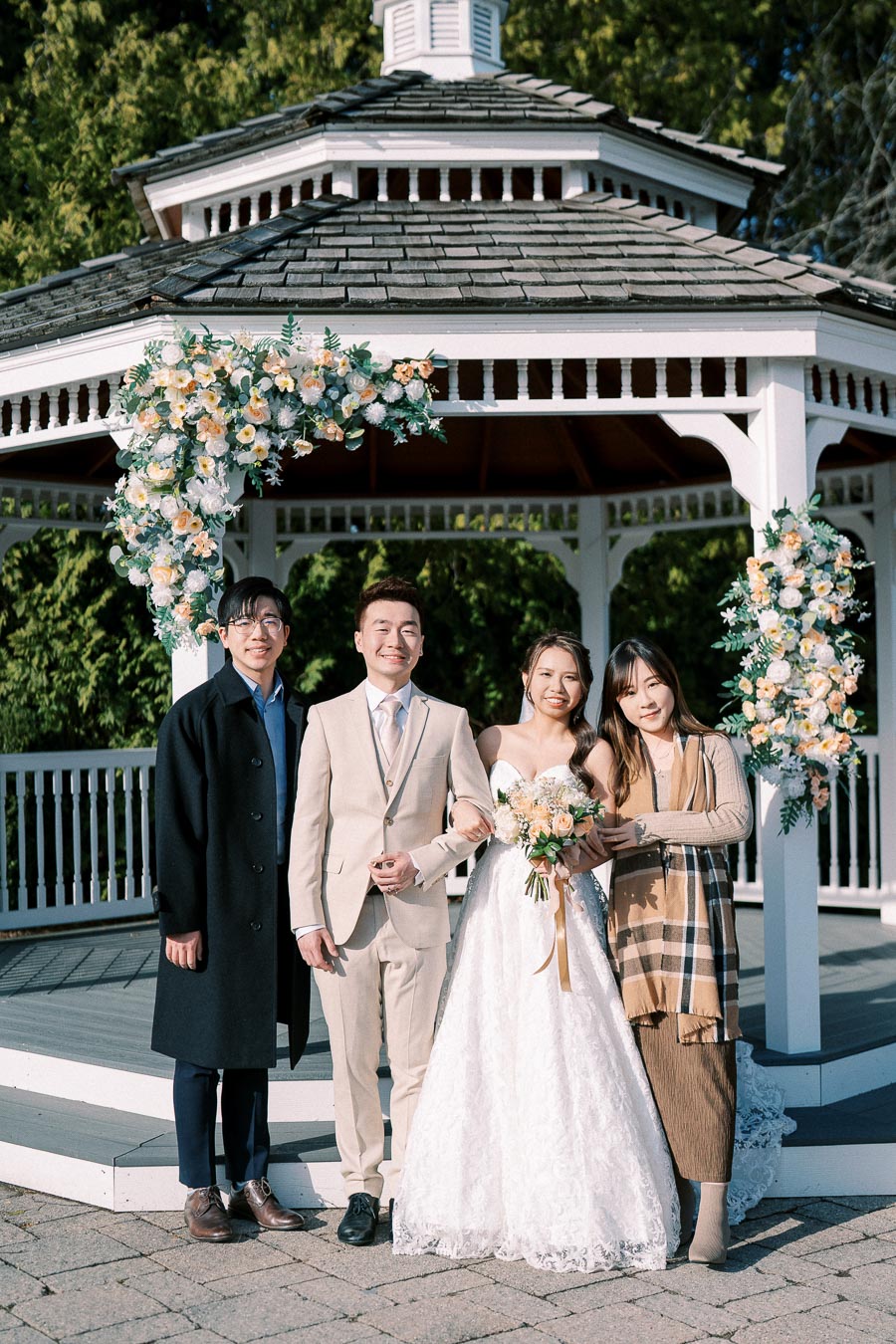 A joyful wedding scene at a gazebo with a newlywed couple and two guests, surrounded by floral decorations and lush greenery.