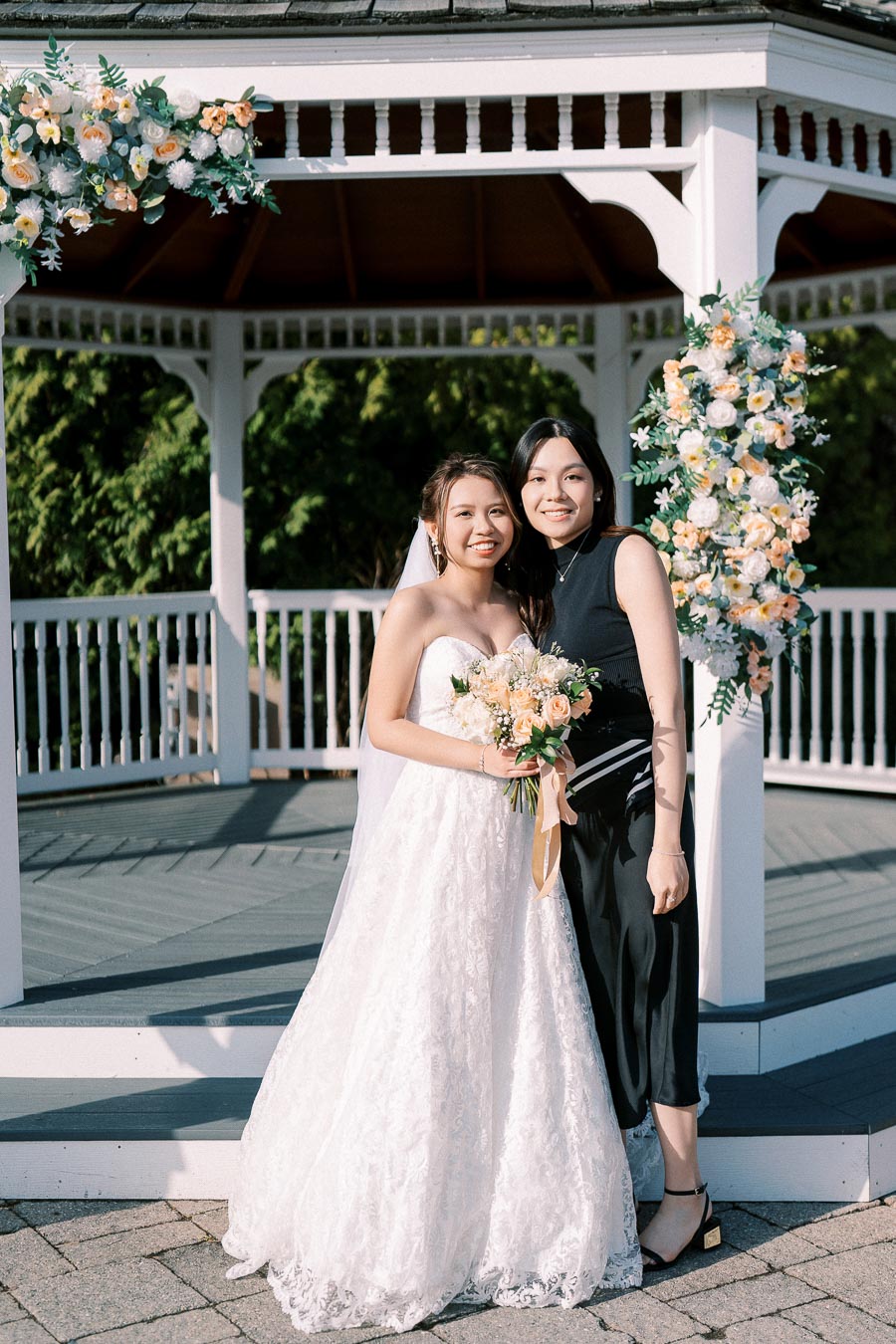 Bride in a white wedding dress stands beside a friend at an outdoor gazebo adorned with floral arrangements, holding a bouquet of cream and peach roses.