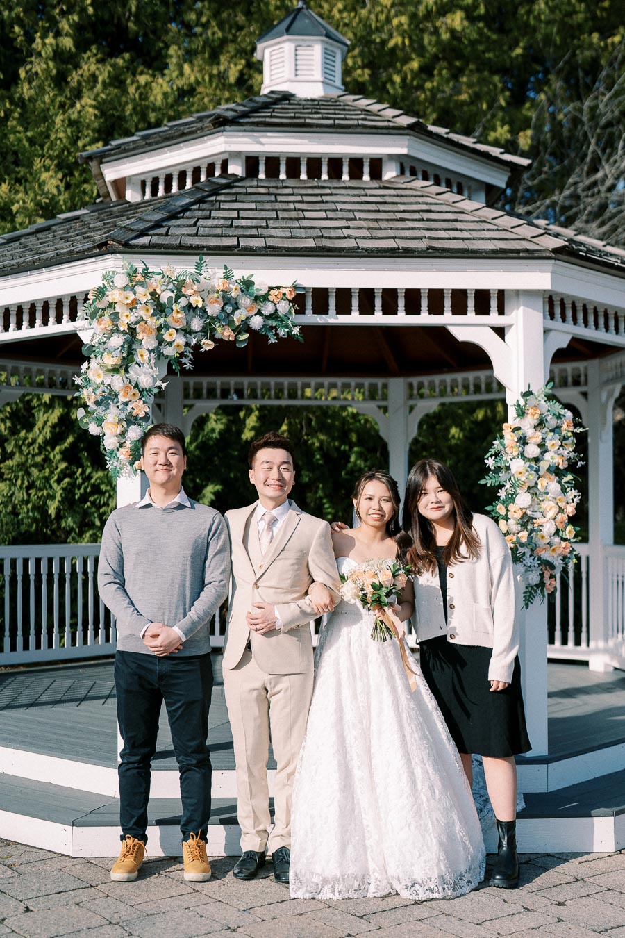 A joyful wedding scene with a newlywed couple and their guests posing in front of a floral-decorated gazebo, set in a lush green garden.