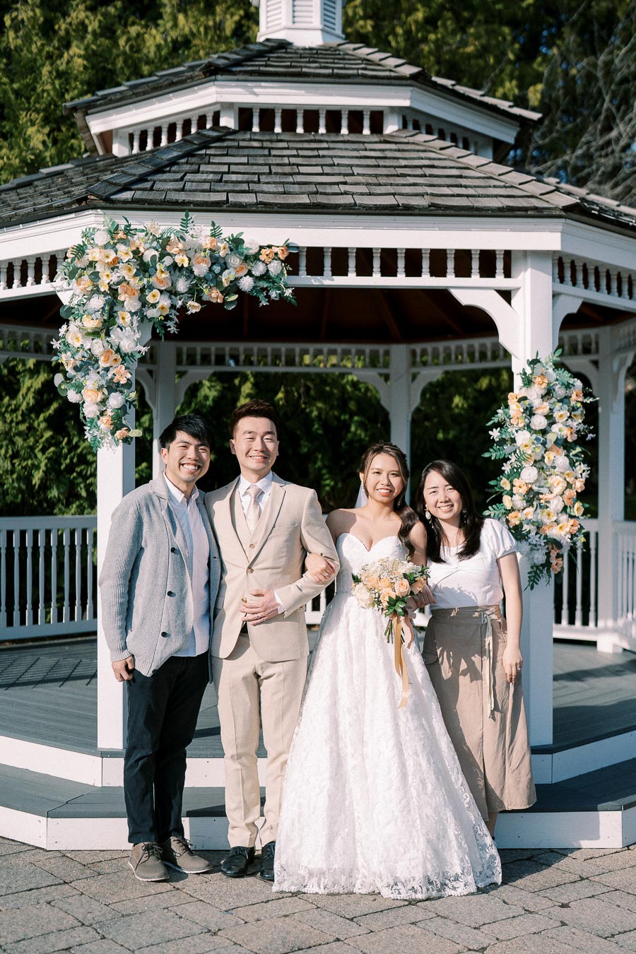 A newlywed couple poses with two guests in front of a beautifully decorated gazebo, adorned with floral arrangements, on a sunny day.
