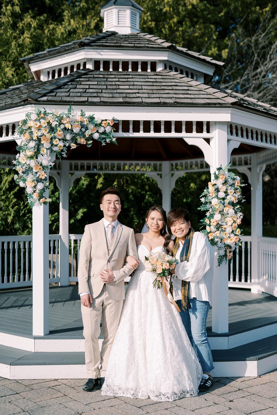 A bride in a white lace wedding dress stands arm-in-arm with a groom in a beige suit and a guest in casual attire. They are posing in front of a white gazebo adorned with floral arrangements on a sunny day.