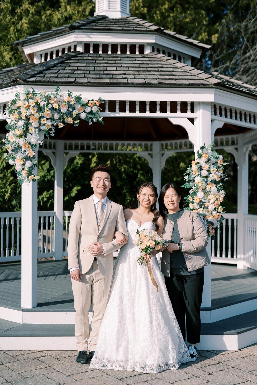 A bride in a white wedding dress stands with a groom in a light beige suit and another person in front of a flower-adorned white gazebo. The sunny outdoor setting and floral decorations suggest a celebratory occasion.