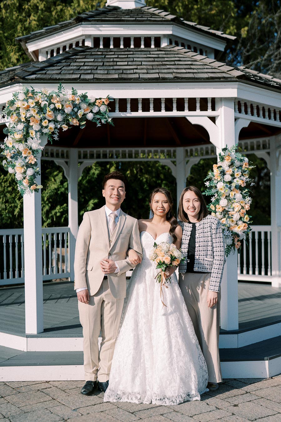 Wedding couple poses with a guest in front of a decorated gazebo with floral arrangements, showcasing a joyful celebration.
