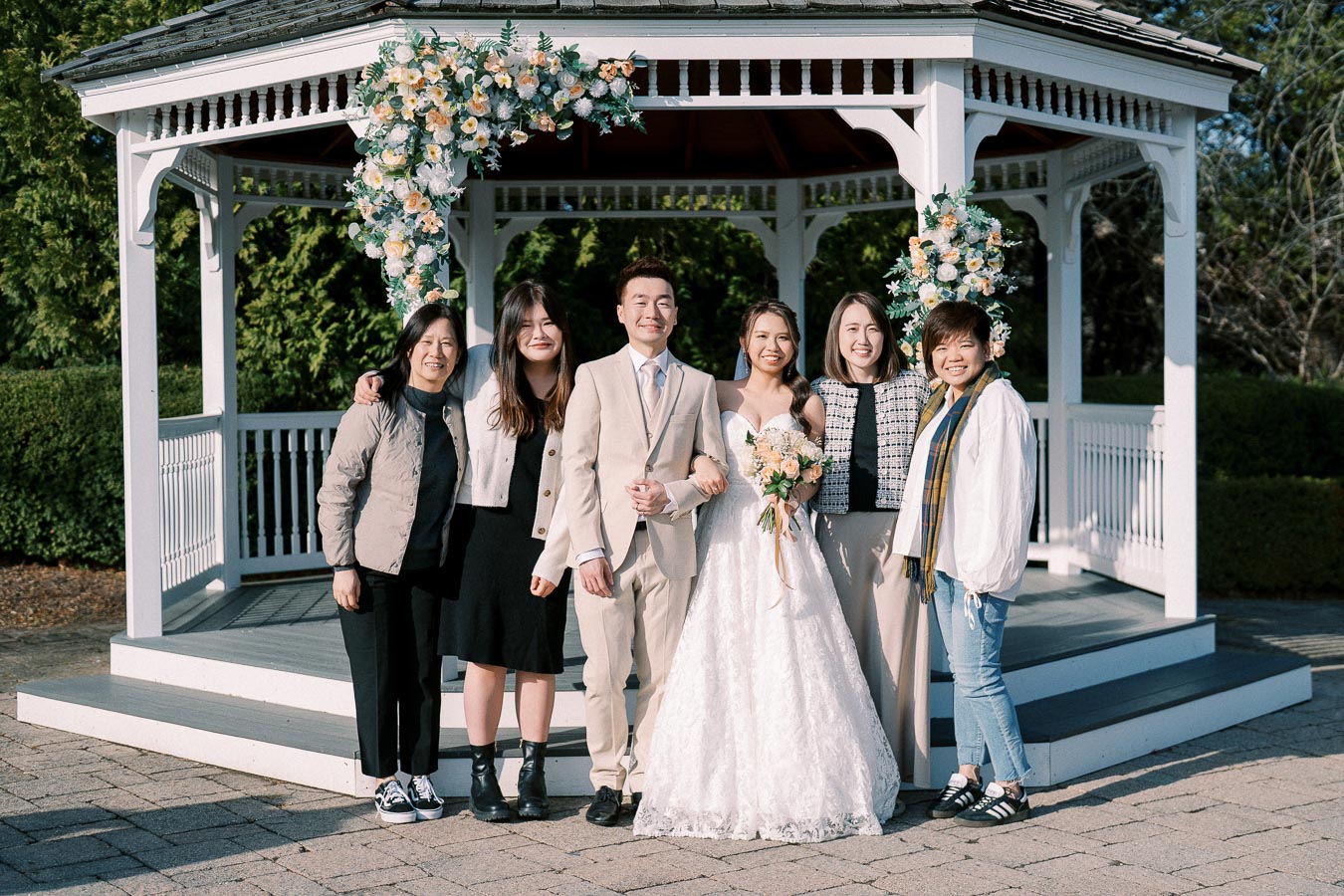 A joyful wedding group posing in front of a beautifully decorated gazebo with floral arrangements, featuring the bride in a white gown holding a bouquet, the groom in a light suit, and four attendees smiling together on a sunny day.