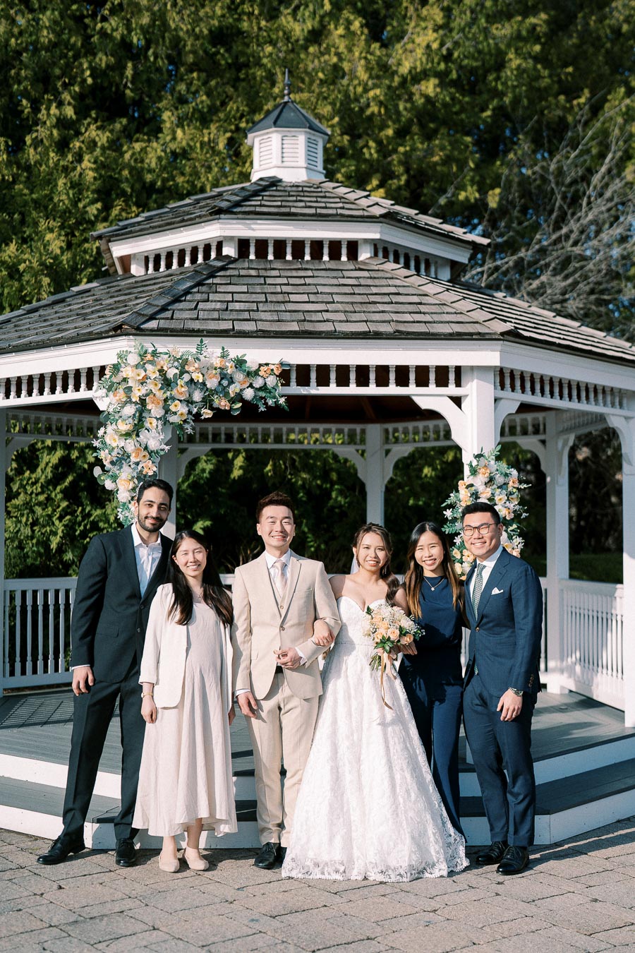 Wedding party posing in front of a decorated gazebo, featuring a bride in a white dress and groom in a beige suit, surrounded by smiling friends in formal attire, with floral accents and lush greenery in the background.