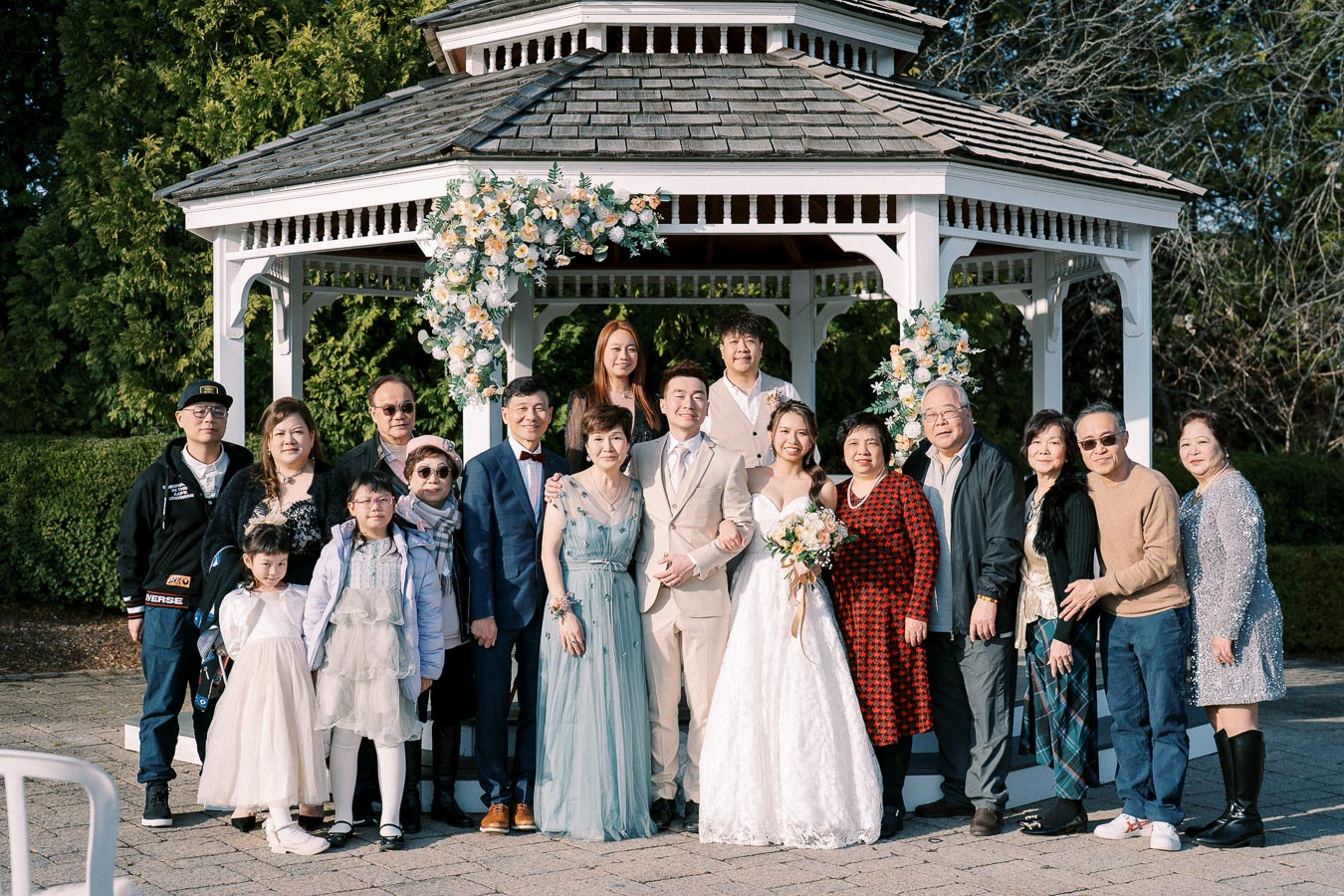 A joyful wedding celebration with a bride and groom surrounded by family and friends in front of a decorated gazebo, capturing a memorable moment in the sunshine.