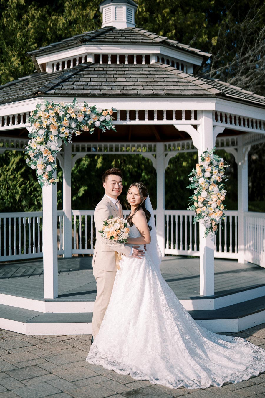 Bride and groom standing in front of a beautifully decorated gazebo adorned with flowers on their wedding day, capturing a romantic moment in natural sunlight.