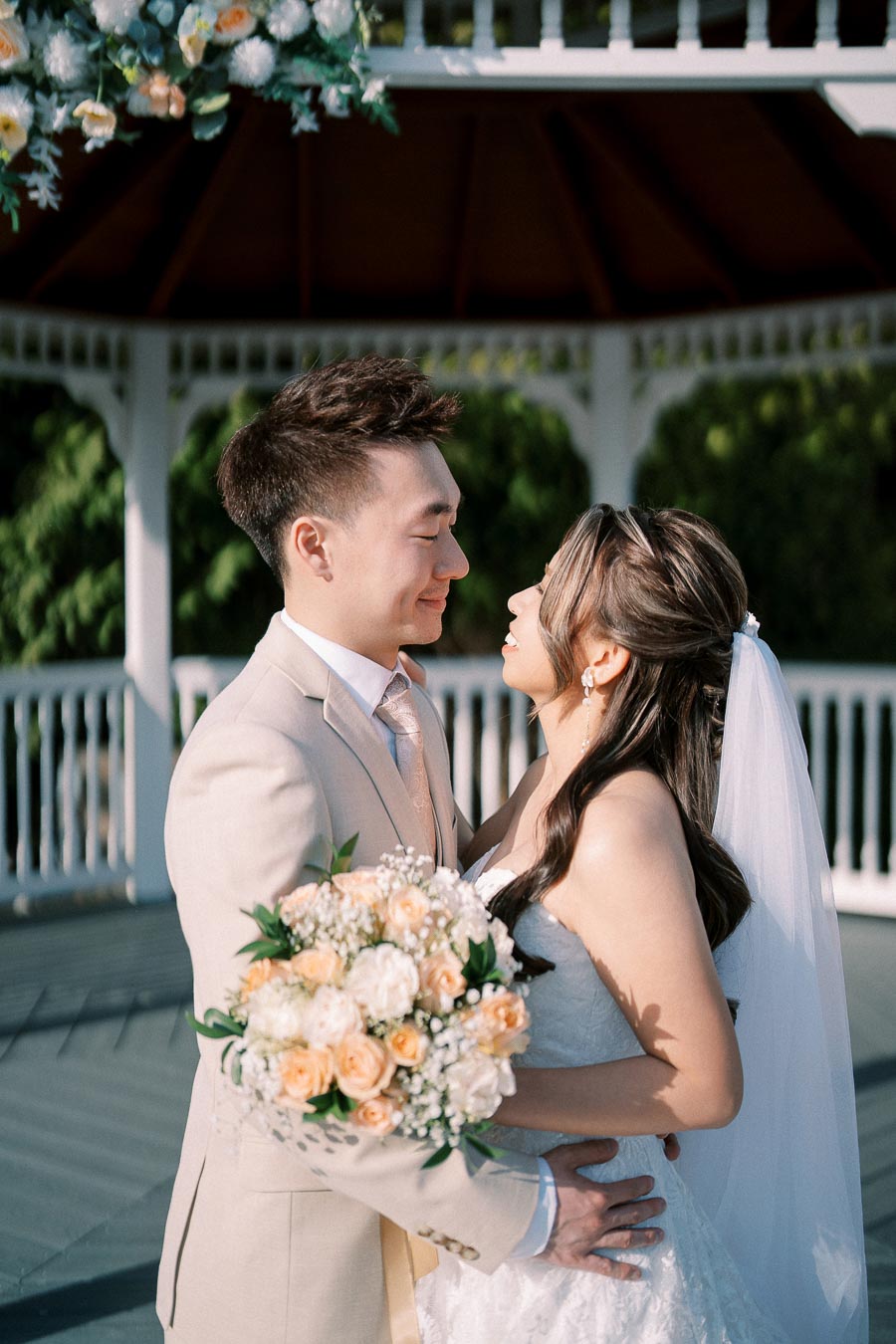 Bride and groom smiling at each other under a decorated gazebo, bride holding a bouquet of peach and white roses, in an outdoor wedding setting.