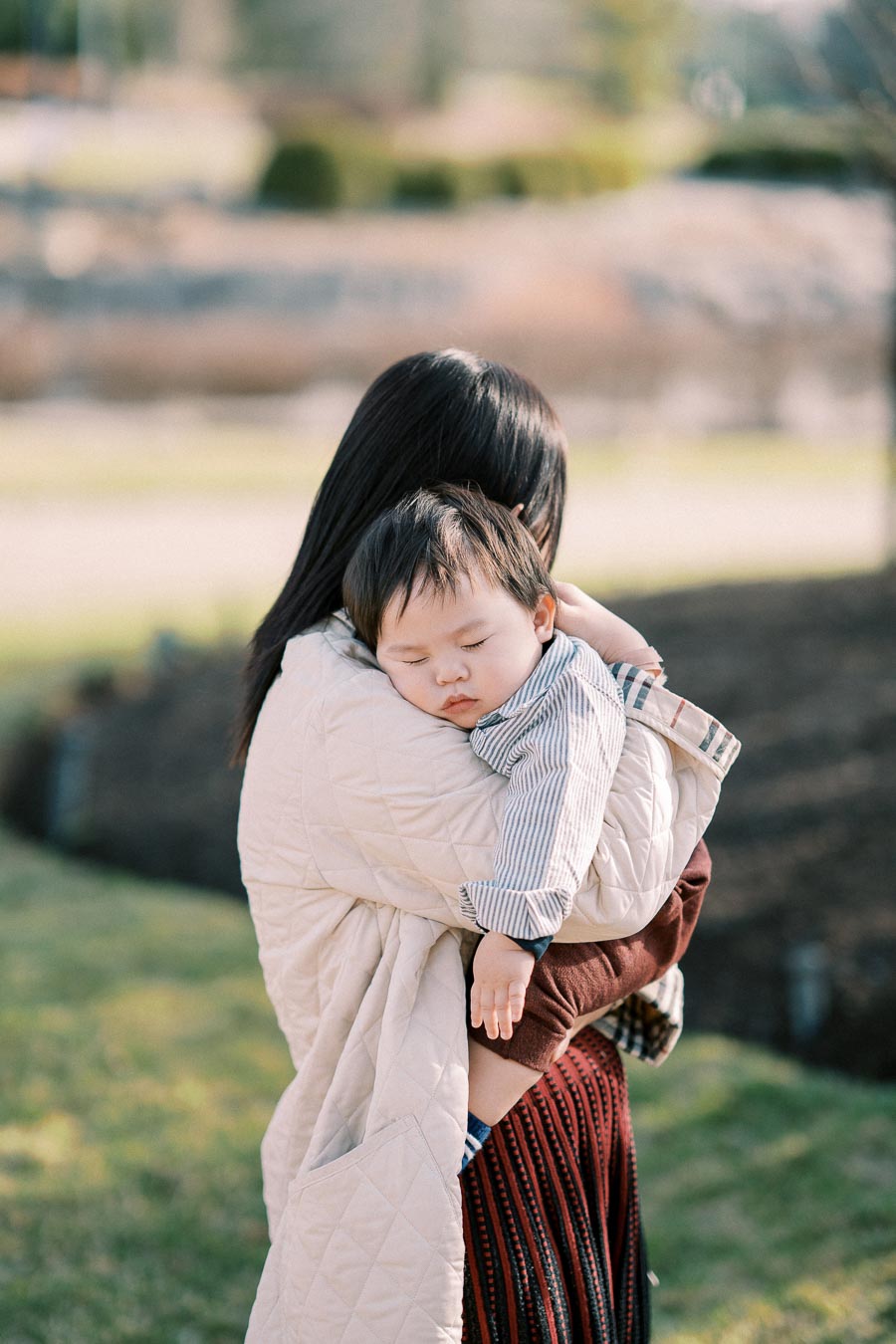 A mother gently holding her sleeping baby outdoors in a serene park setting, conveying warmth and tenderness.