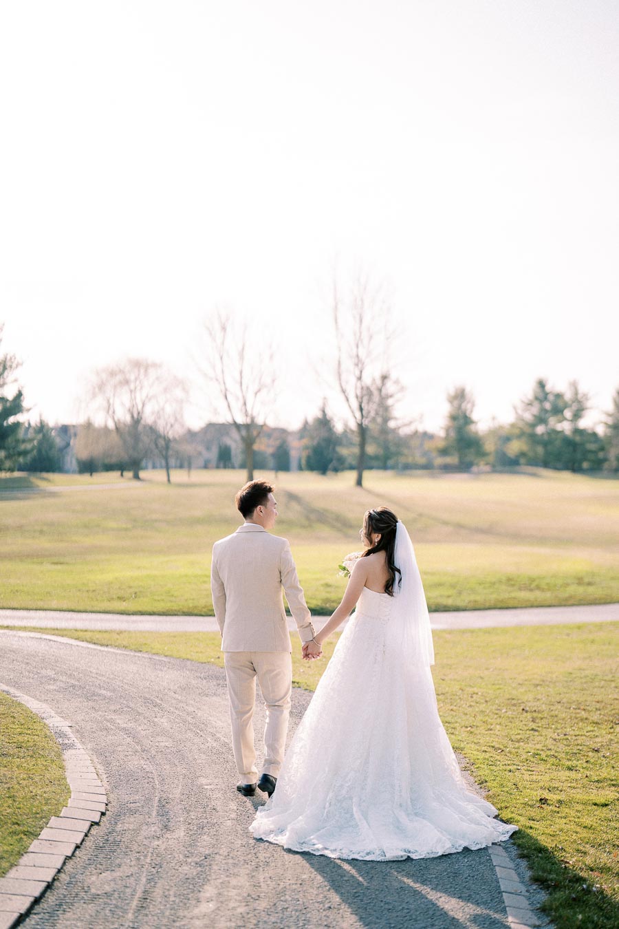 Couple holding hands walking down a path in a peaceful park setting, bride in a white gown and groom in a beige suit, with lush green grass and trees in the background on a sunny day.