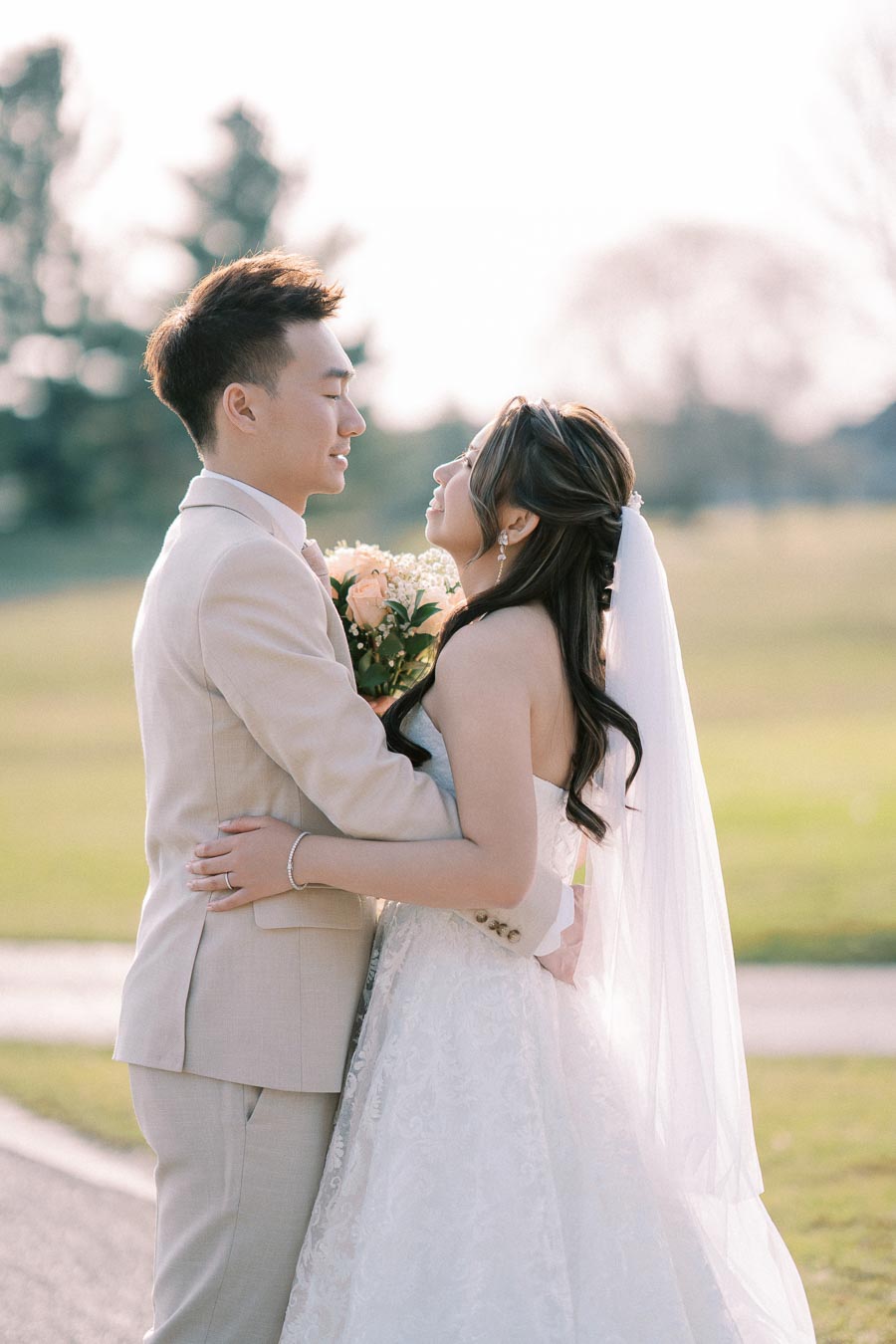 A bride and groom embrace outdoors during a sunny wedding day, surrounded by lush greenery. The bride wears a white lace gown and veil, holding a bouquet of pale roses, while the groom is dressed in a light-colored suit.